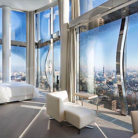 The Westin Hamburg Elbphilharmonie Hotel Екстериор снимка The photo shows a modern hotel room with a stunning, panoramic view through large, uniquely designed windows. The room features a sleek, minimalist aesthetic with light-colored furnishings, including a white bed and a couple of chairs. The walls are 