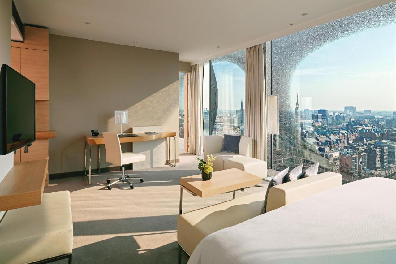The Westin Hamburg Elbphilharmonie Hotel Екстериор снимка The photo depicts a modern hotel room featuring a contemporary design. The space includes a large window that offers a panoramic view of the city beyond. Inside, there are stylish furniture elements such as a small dining table and chairs, a desk wit