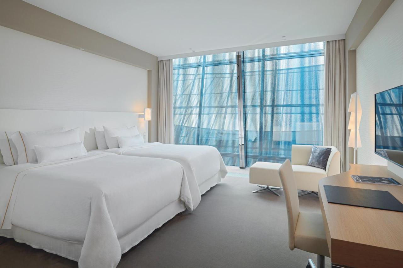 The Westin Hamburg Elbphilharmonie Hotel Екстериор снимка The photo shows a modern hotel room featuring two white beds with plush bedding. There is a large window with sheer curtains that allows natural light to filter in, offering a view outside. The room has neutral-colored walls and carpeting, creating a