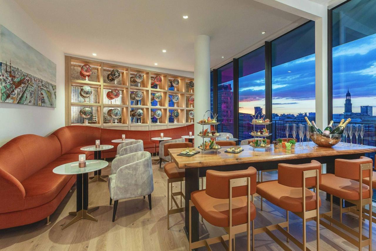 The Westin Hamburg Elbphilharmonie Hotel Екстериор снимка The photo shows a modern and stylish restaurant or café interior. It features a combination of comfortable seating, including a curved, orange sofa and gray chairs. The tables are round with light-colored tops, and there are several tiered stands dis