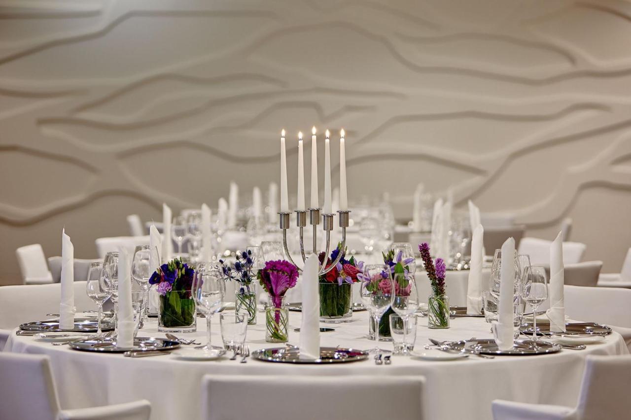 The Westin Hamburg Elbphilharmonie Hotel Екстериор снимка The photo depicts an elegantly set round dining table. In the center, there is a candelabra with several tall candles, providing a soft, warm light. Surrounding the candelabra are small glass vases filled with colorful flowers, adding a vibrant touch