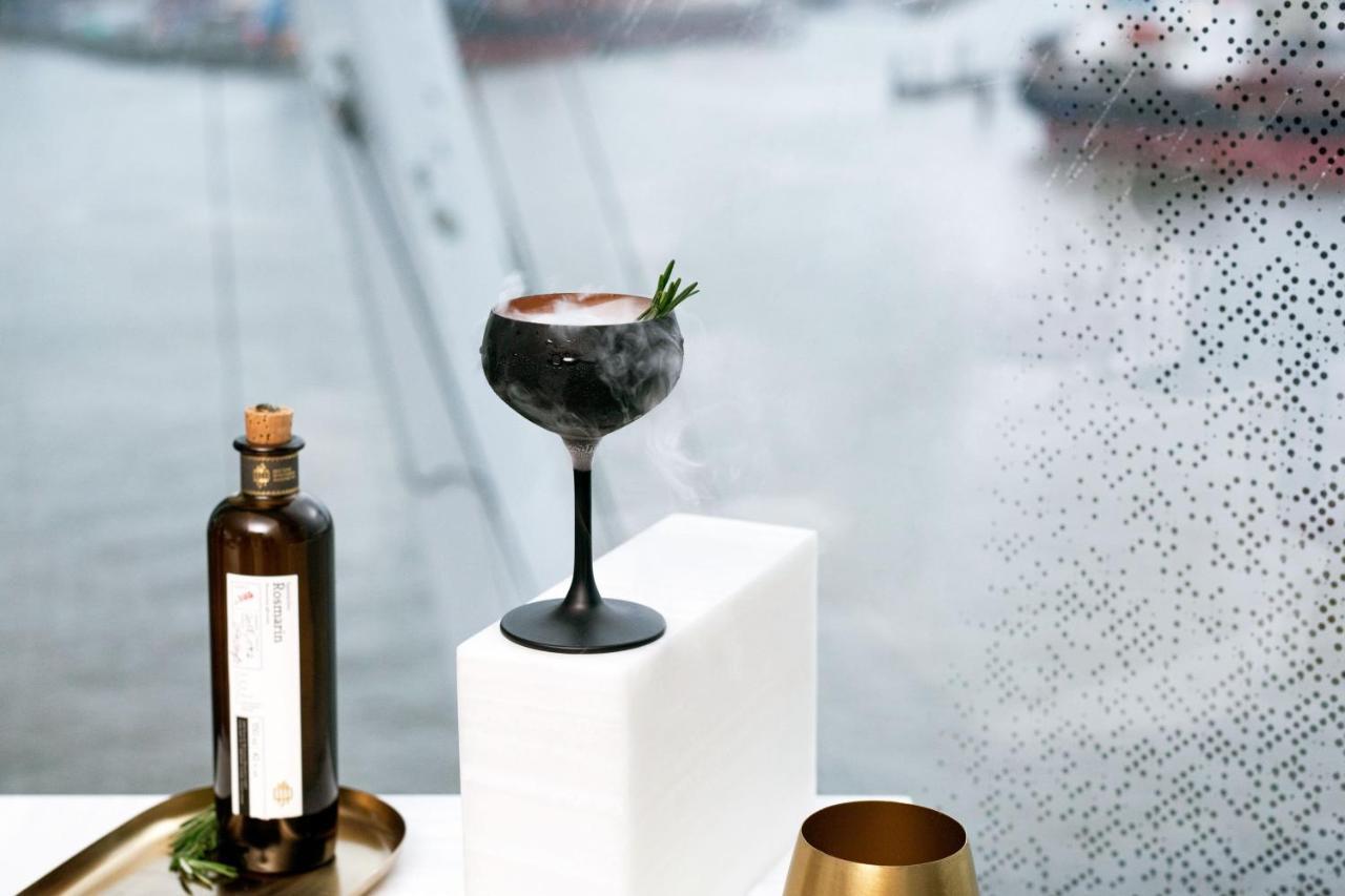 The Westin Hamburg Elbphilharmonie Hotel Екстериор снимка The photo features a stylish cocktail elegantly presented on a white pedestal. The cocktail is served in a sophisticated black stem glass, topped with a garnish of rosemary. Beside it, there is a dark glass bottle, likely containing a spirit or mixer