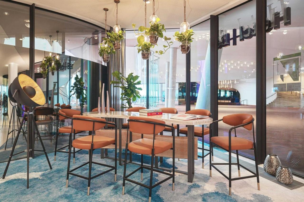 The Westin Hamburg Elbphilharmonie Hotel Екстериор снимка The image depicts a modern indoor seating area designed for gatherings or meetings. There is a long table surrounded by several bar-style chairs with brown upholstery. Above the table, there are hanging plants that add a touch of greenery to the spac