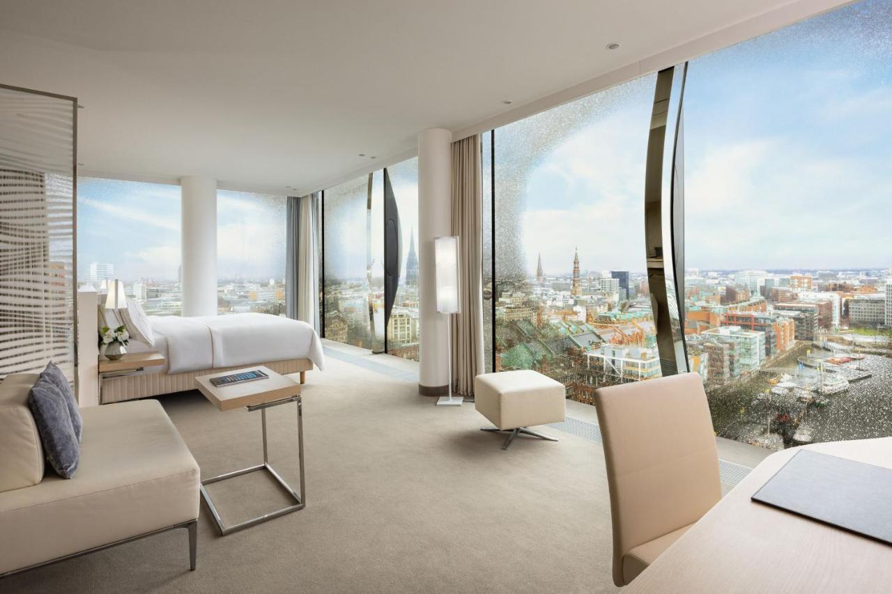The Westin Hamburg Elbphilharmonie Hotel Екстериор снимка The photo shows a modern hotel room with large floor-to-ceiling windows that provide a panoramic view of a city landscape. The room features a minimalist design with neutral tones. There's a king-sized bed positioned in the center, a sleek desk and c